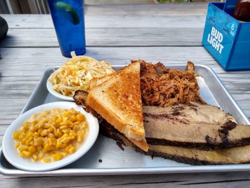 A metal tray with sliced brisket, pulled pork, coleslaw, corn, and toasted bread, alongside a blue drink.