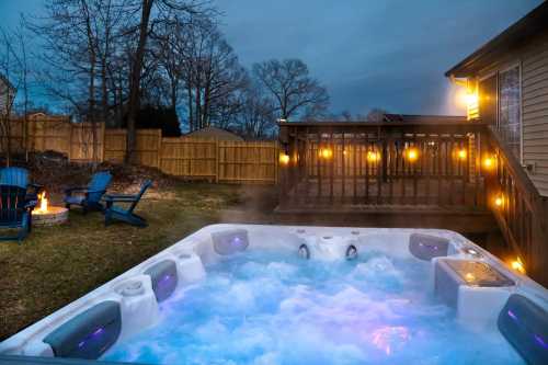 A hot tub with blue water and lights, surrounded by a wooden deck and a cozy fire pit in a backyard at dusk.