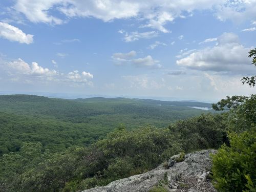 A panoramic view of lush green mountains under a partly cloudy sky, with a distant lake visible in the background.