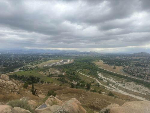 A panoramic view of a green valley surrounded by hills under a cloudy sky, with a winding road in the foreground.