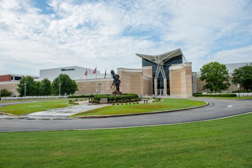 A modern building with a large statue in front, surrounded by green grass and trees under a cloudy sky.