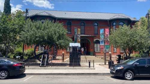 Historic red brick building with banners, surrounded by trees and parked cars, under a clear blue sky.