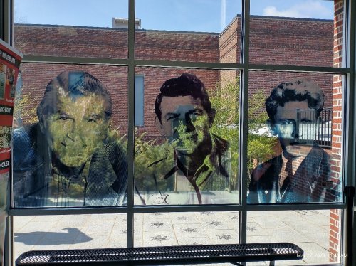 Three large, artistic portraits of men are displayed on a window, with a brick building and trees visible outside.