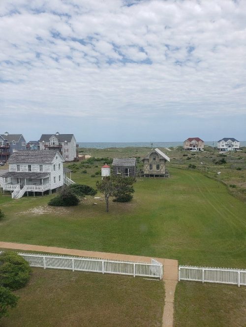 Coastal scene featuring a grassy area, two houses, a lighthouse, and the ocean under a cloudy sky.