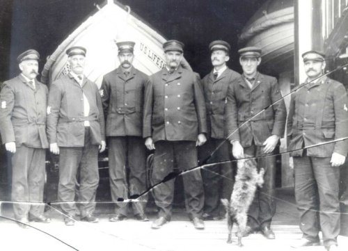 A historic black-and-white photo of eight uniformed men, standing together with a dog, near lifeboats.