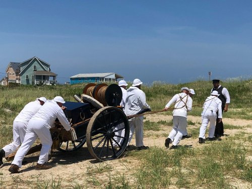 A group of people in white clothing pulls a historical cannon on a sandy path, with houses and grass in the background.