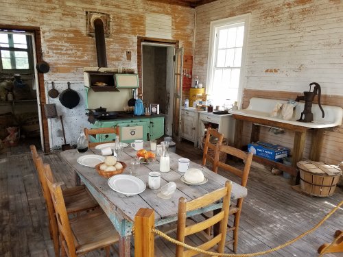 A rustic kitchen with a wooden table set for a meal, featuring an old stove, sink, and vintage decor.