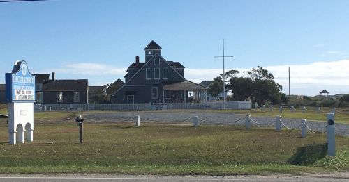A historic building with a gray exterior and a white fence, set against a clear blue sky and grassy landscape.