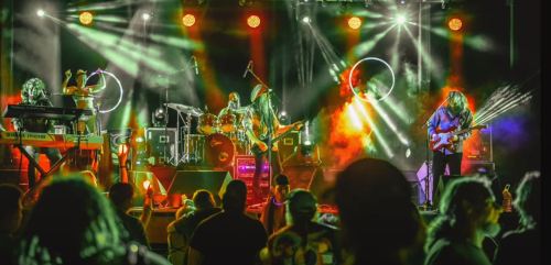 A lively band performs on stage with colorful lights, while an engaged audience watches from the front.