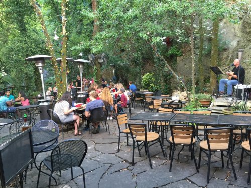 A lively outdoor café with diners at tables, surrounded by greenery, and a musician performing on stage.