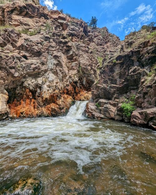 A waterfall cascades into a rocky pool, surrounded by steep cliffs and greenery under a blue sky.