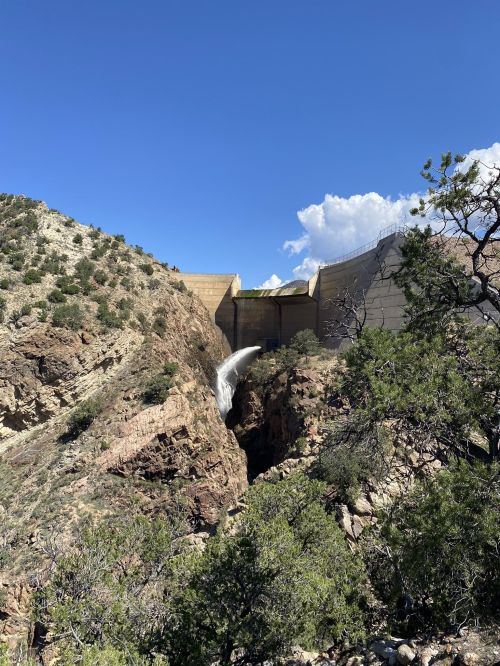 A dam releases water into a rocky canyon, surrounded by greenery and under a clear blue sky with scattered clouds.