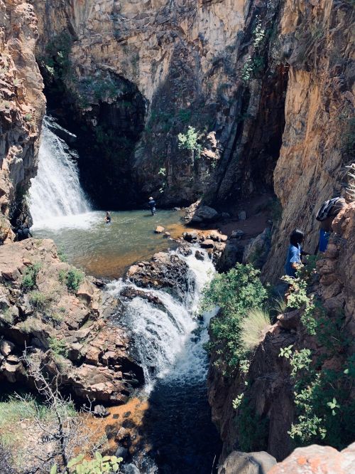 A scenic waterfall cascades into a pool surrounded by rocky cliffs and greenery, with people enjoying the water.