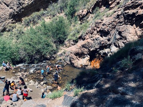 People gather by a rocky stream surrounded by greenery, enjoying a sunny day outdoors.