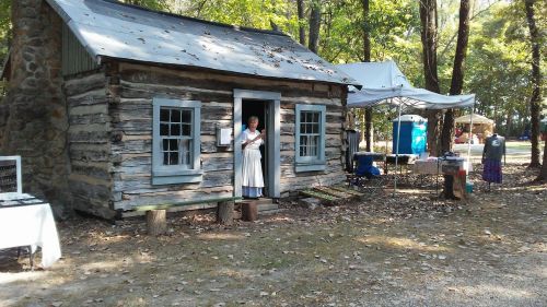 A woman in period clothing stands at the door of a rustic log cabin, surrounded by trees and vendor tents.