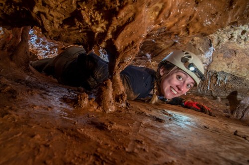 A person in a helmet crawls through a narrow cave passage, smiling amidst rocky, muddy surroundings.