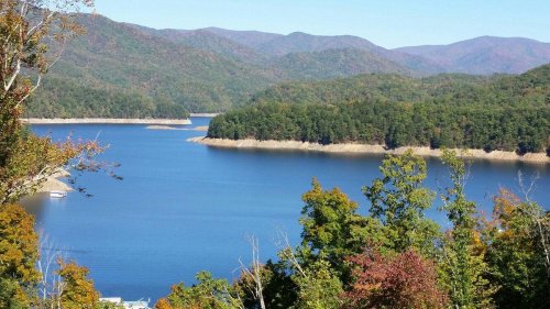 A serene lake surrounded by lush green trees and distant mountains under a clear blue sky.