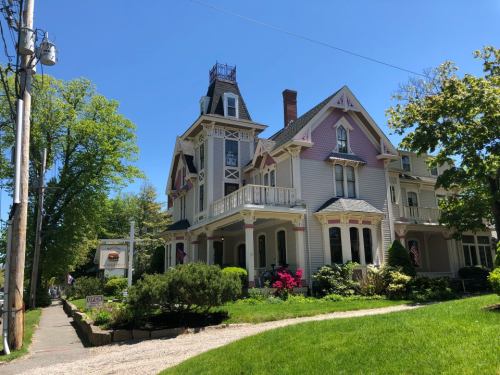 A large, ornate Victorian house with a tower, surrounded by greenery and colorful flowers on a sunny day.