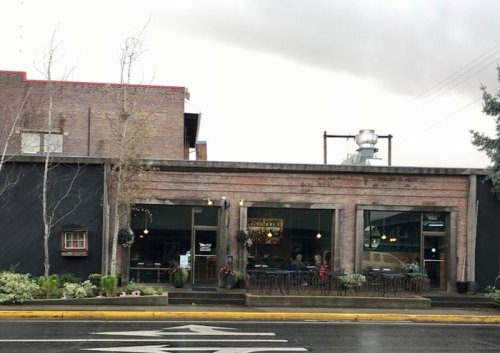 A brick building with large windows, plants outside, and a sign, set against a cloudy sky.