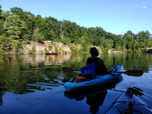 A person in a kayak paddles on a calm lake, surrounded by lush greenery and rocky cliffs in the background.