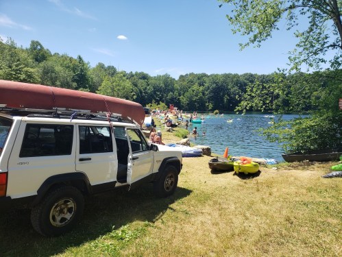 A white SUV parked by a lake, with people enjoying the water and greenery in the background on a sunny day.