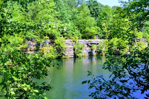 Lush green trees surround a calm, reflective body of water with rocky cliffs in the background.