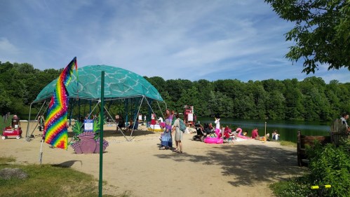 A sandy beach area with a colorful dome structure, people relaxing, and a lake surrounded by green trees.