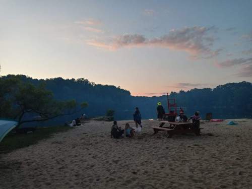 A sandy beach at dusk with people gathered, trees in the background, and a calm lake reflecting the sky.