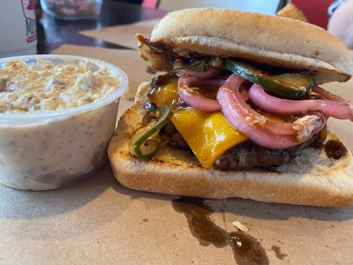 A close-up of a cheeseburger with pickles and onions, alongside a cup of creamy coleslaw on a paper surface.