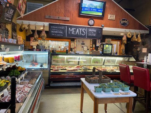 Interior of a butcher shop featuring meat displays, fresh produce, and a chalkboard menu in a rustic setting.