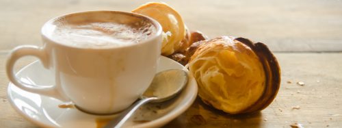 A cup of coffee on a saucer next to flaky croissants on a wooden table.
