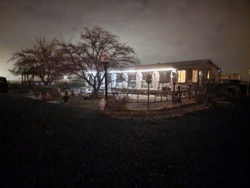 A dimly lit house at night, surrounded by trees and a fence, with chairs and tables visible outside.