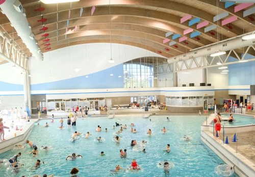 A busy indoor pool with people swimming and relaxing on floats under a high, colorful ceiling.