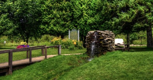 A serene park scene featuring a stone fountain, lush greenery, and a wooden bridge leading to a sign.