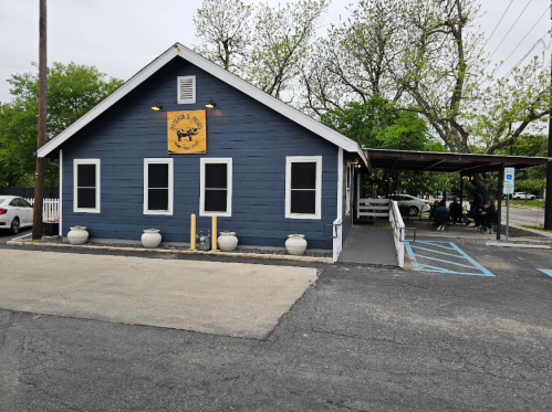 A blue wooden building with a sign, surrounded by trees and parked cars, featuring outdoor seating.