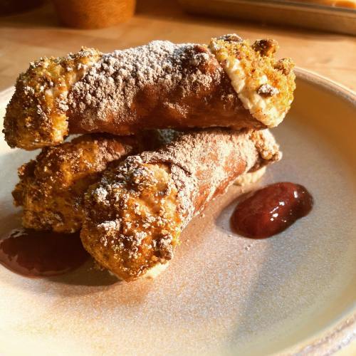 Three cannoli dusted with powdered sugar, served on a plate with a dollop of red fruit sauce.