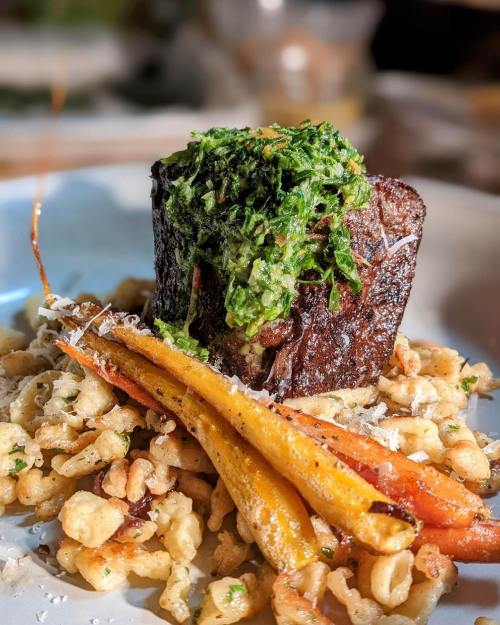 A beautifully plated dish featuring a tender piece of meat topped with green sauce, served with crispy pasta and colorful carrots.