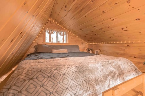Cozy attic bedroom with wooden walls, a large bed, and soft lighting from a window.