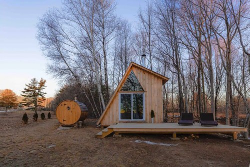 A modern wooden cabin with an angled roof, deck, and a barrel sauna, surrounded by trees in a serene landscape.