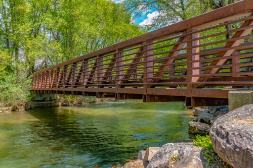 A wooden bridge spans a clear stream, surrounded by lush green trees and rocky banks under a bright blue sky.