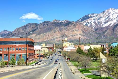 A scenic view of a city street with mountains in the background, featuring buildings and clear blue skies.