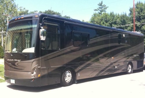 A sleek, dark-colored RV parked on a sunny street, surrounded by trees.