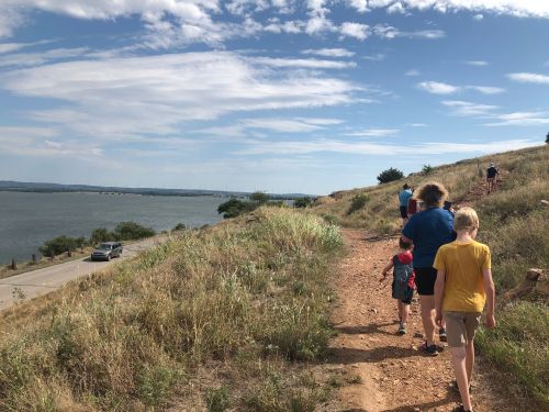A group of children walking along a dirt path by a lake, with grassy hills and a blue sky in the background.