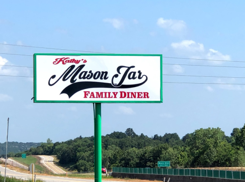 Sign for "Kathy's Mason Jar Family Diner" with a green pole, set against a blue sky and green landscape.