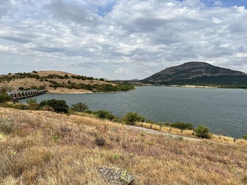 A scenic view of a lake surrounded by hills and a cloudy sky, with dry grass in the foreground.