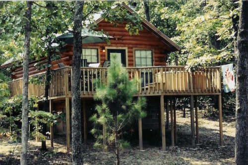 A rustic log cabin on stilts surrounded by trees, featuring a deck with chairs and a colorful flag hanging.