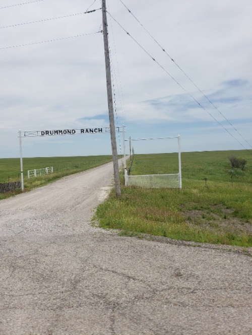 A gravel road leads to a white gate marked "Drummond Ranch," surrounded by green fields under a cloudy sky.