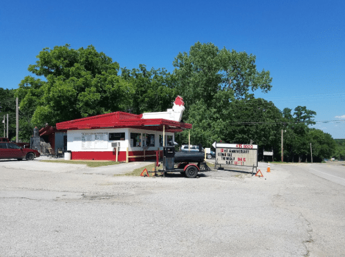 A small roadside diner with a red roof, surrounded by trees, featuring a sign for an anniversary event.