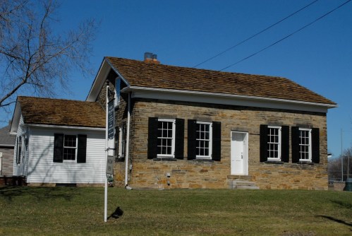 A stone and wood house with a sloped roof, featuring black shutters and a white door, set against a clear blue sky.