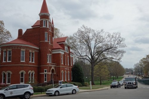 Historic red brick building with a pointed tower, surrounded by trees and a street with parked cars on a cloudy day.